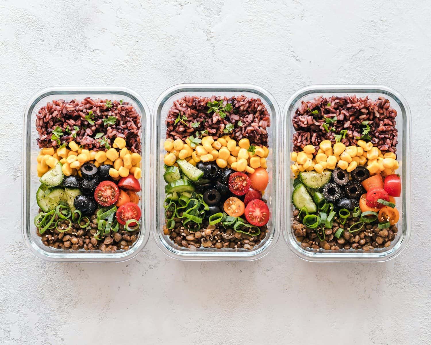 Three glass containers filled with rice, corn, olives, tomatoes, cucumbers, and lentils