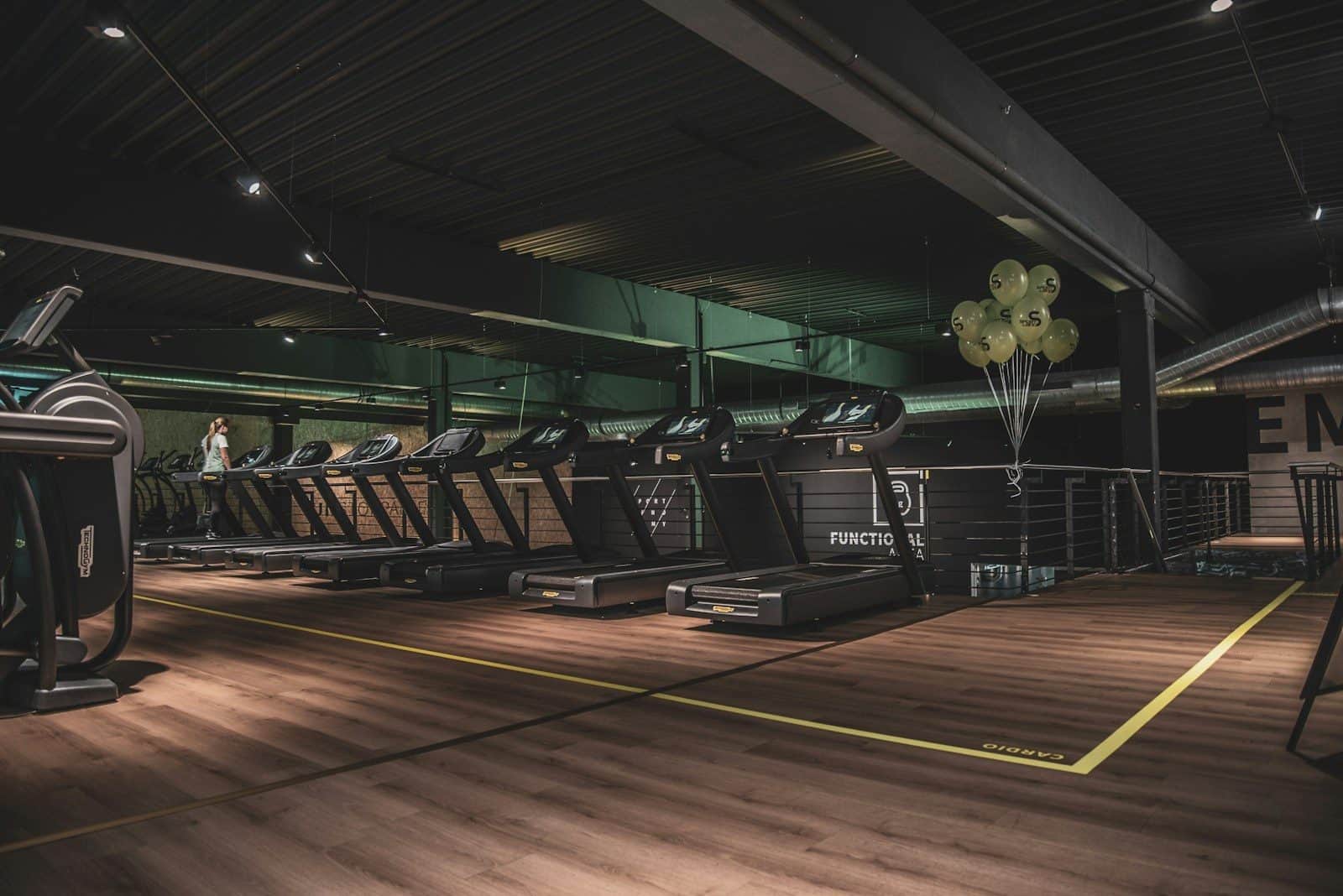 A row of treadmills in a gym with wooden floors and green balloons