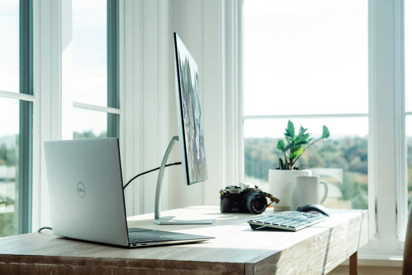A laptop, monitor, camera, and plant on a wooden desk in a sunlit office