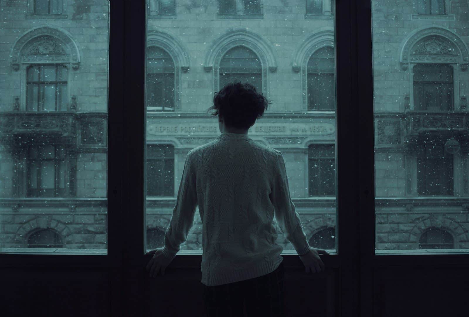 A person in a white sweater looking out a window at falling snow