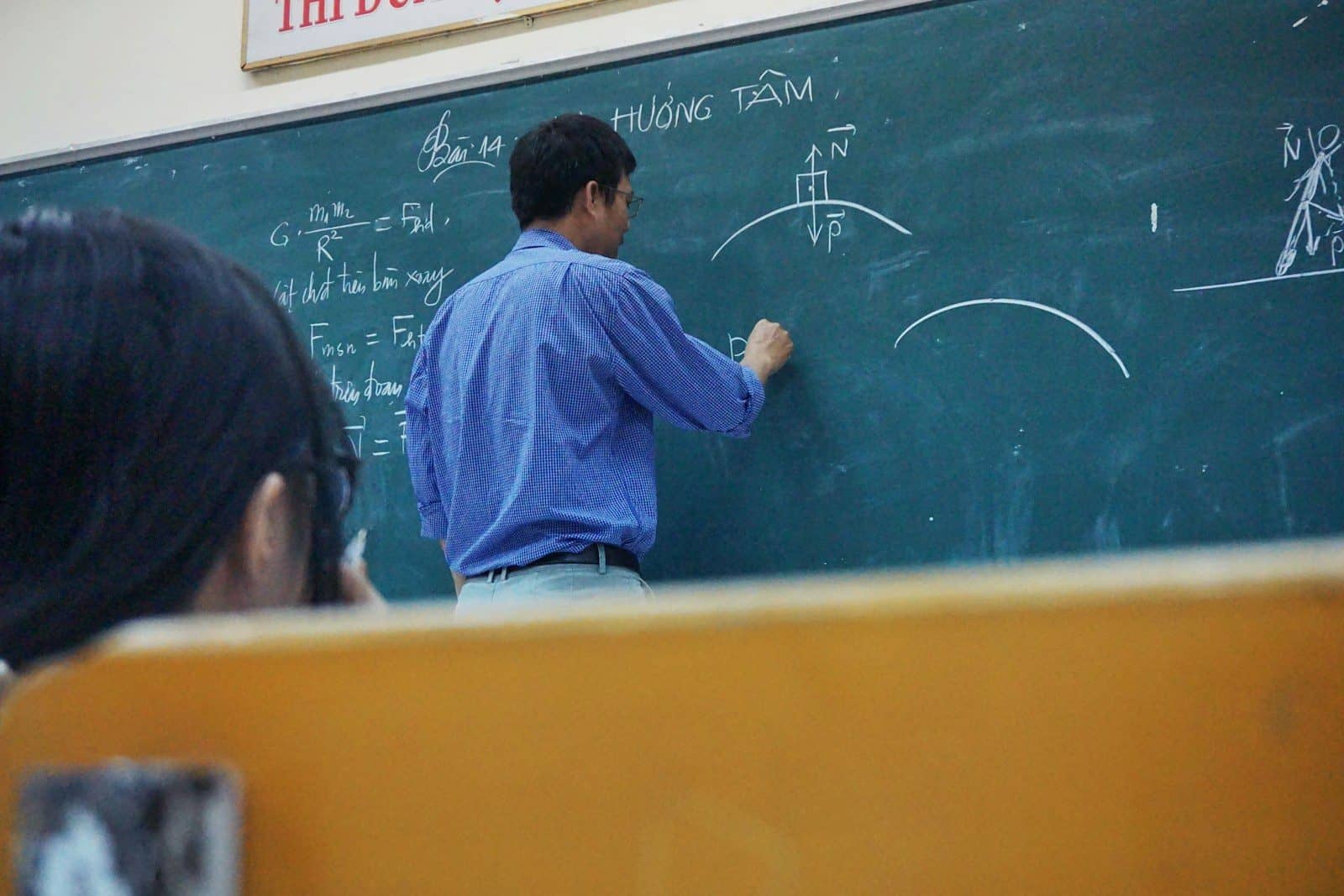A teacher writing physics equations on a green chalkboard in a classroom