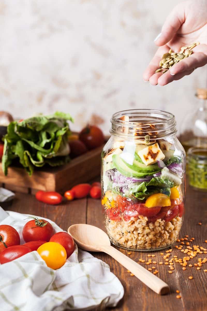 A hand sprinkling pumpkin seeds into a layered salad in a glass jar