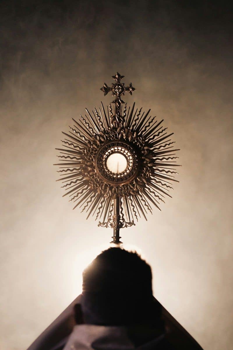 A person holding a golden monstrance with a cross against a soft light