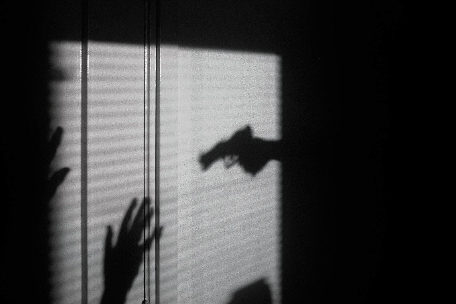 Shadows of hands and a handgun cast against a window with closed blinds