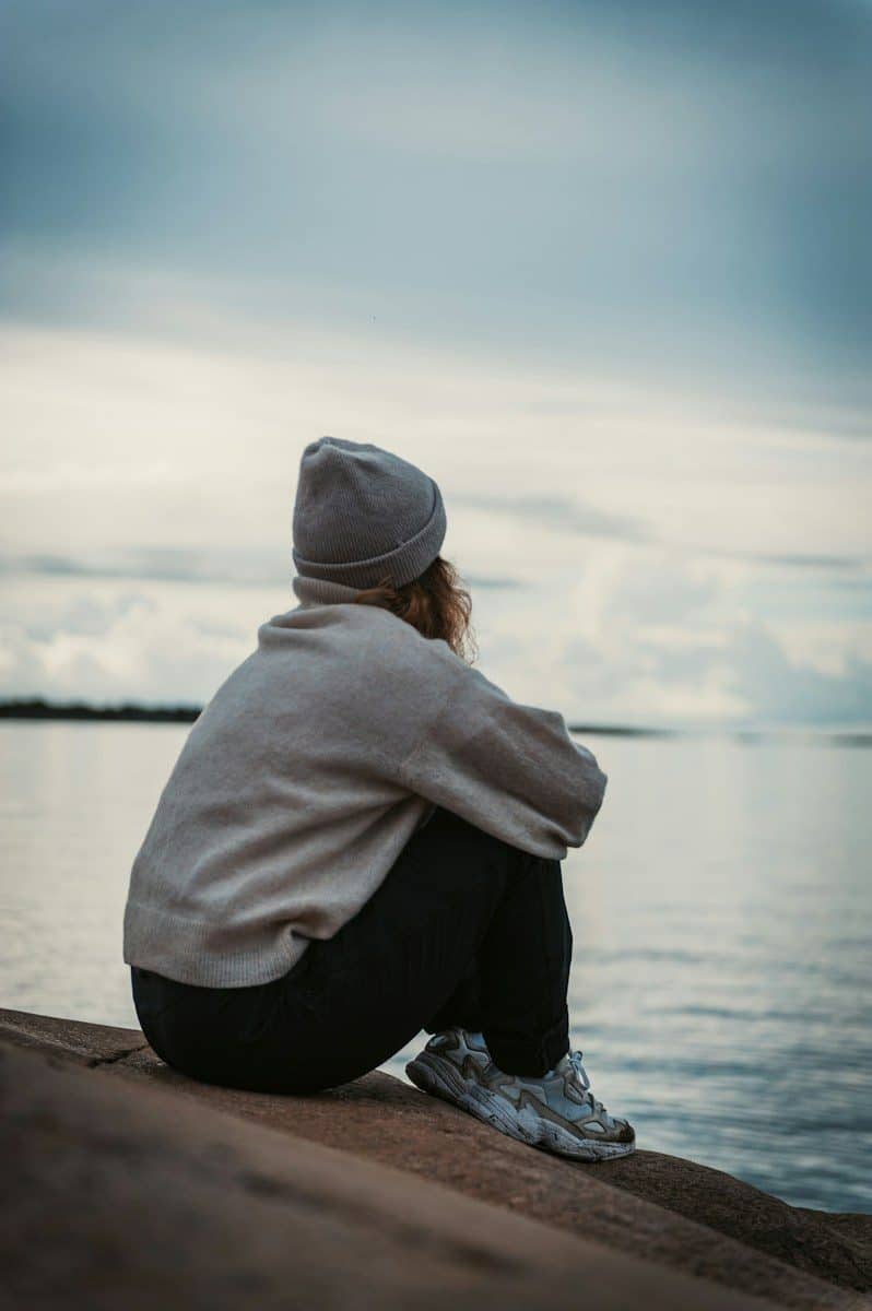 A person in a beanie and sweater sitting on a rock by the water